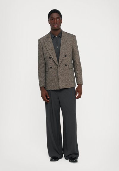 Man i en strukturerad dubbelknäppt blazer, mörk zip-up skjorta, vida byxor och svarta skor, som står mot en enkel vit bakgrund.