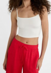 Weißes, kurzes Tanktop mit dünnen Trägern, kombiniert mit roten Leinen-Hosen mit Kordelzug. Glatte Struktur, schlichtes Design und lebendiger Farbkontrast.
