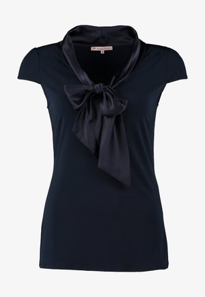 Kobieta w czarnej bluzce z krótkim rękawem z dużą kokardą, niebieskich obcisłych dżinsach, czarnych botkach za kostkę, trzyma czarną kopertówkę, idzie do przodu.