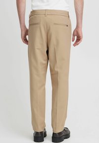 Beige trousers with a relaxed fit, elastic waistband, and two back pockets featuring horizontal zippers. Smooth texture and minimal design.