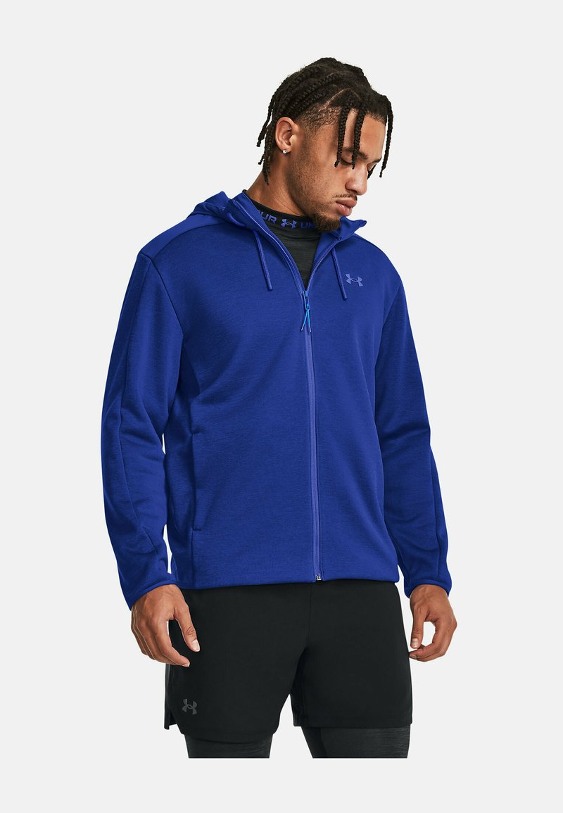Blaue Kapuzenjacke mit Reißverschluss, verstellbarem Kapuzenzug, langen Ärmeln und seitlichen Taschen. Hat eine glatte Textur und ein Logo auf der Brust. Kombiniert mit schwarzen Shorts.