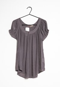 Blusa grigia a maniche corte con tessuto arricciato, scollatura a girocollo, due bottoni al colletto, appesa a una gruccia nera.