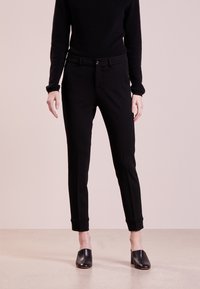 Schwarze, tapered Hosen mit glatter Textur, ausgestattet mit einer figurbetonten Taille und umgeschlagenen Säumen. Getragen mit einem schwarzen Pullover und Slippern.