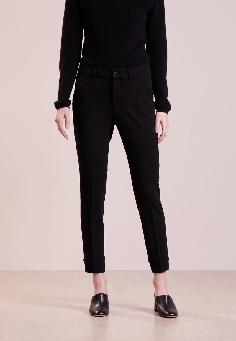 Schwarze, tapered Hosen mit glatter Textur, ausgestattet mit einer figurbetonten Taille und umgeschlagenen Säumen. Getragen mit einem schwarzen Pullover und Slippern.