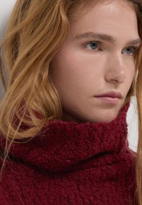 Junge Person mit langen, welligen roten Haaren, die einen dicken, strukturierten, dunkelroten Rollkragenpullover trägt, nach rechts blickend mit neutralem Ausdruck.