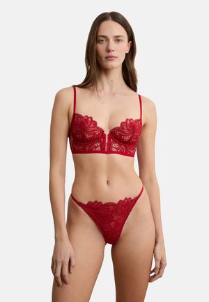 Donna che indossa un reggiseno di pizzo floreale rosso con chiusura frontale e perizoma di pizzo abbinato, in piedi davanti a uno sfondo bianco semplice.