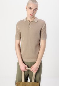 Beige polo tričko s krátkým rukávem a texturovaným vzorem, s knoflíkovým límcem a kontrastním lemováním rukávů. Zelené kalhoty a béžová taška.