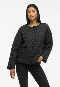 Juoda siuvinėta striukė su širdelių raštu, apvaliu iškirpimu, spaudžiamu užsegimu ir dviem šoninėmis kišenėmis, vilkima ant tamsių džinsų.