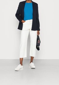 Veste de blazer marine, haut turquoise, jean blanc raccourci avec ourlet effiloché et baskets blanches avec accent marine. Sac à main noir avec bandoulière.