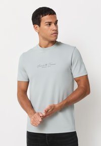 Világosszürke, rövid ujjú póló, puha pamutkeverék anyagból, finom hímzett szövegmintával a mellkason.