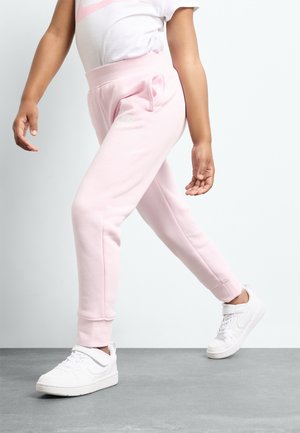 Barn iført lyserøde joggingbukser og hvide sneakers, stående med det ene ben bøjet og armene afslappet ned langs siderne.
