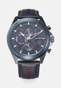 Montre pour homme avec un boîtier rond en métal bleu marine, un cadran noir texturé, des aiguilles argentées et un bracelet en cuir sombre avec des coutures rouges.