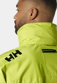 Chaqueta impermeable verde lima con cuello alto, logo negro y franjas reflectantes. Textura suave con un hardware mínimo.