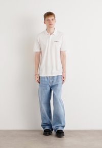 Hvit polo skjorte med en liten svart "HUGO"-logo, kombinert med lyseblå vide jeans og svarte slip-on sko. Enkelt, avslappet antrekk.