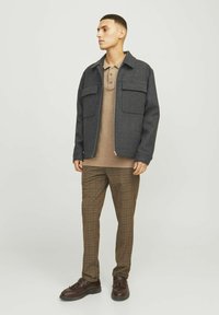 Veste à carreaux zippée en gris foncé, associée à un polo brun et à un pantalon à carreaux bruns. Des chaussures brunes complètent la tenue.