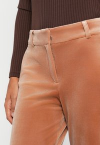 Primer plano de una persona que lleva una camiseta de manga larga de canalé marrón y pantalones de terciopelo color melocotón de tiro alto con cremallera y trabillas para cinturón.
