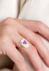 Goldring am Finger mit rechteckiger Fläche, lila Herz und kleineren Formen, umgeben von kleinen klaren Steinen.