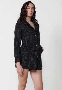 Rochie din tweed negru cu un model în carouri, cu margini destrămate și guler. Prezintă detalii cu nasturi și buzunare laterale.