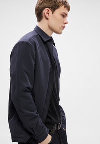 Lahka mornarsko modra jakna z ovratnikom, vidnimi šivi in gladko teksturo, nošena preko črne majice, kombinirana s temnimi kavbojkami.