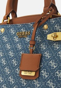 Torebka z dżinsu z powtarzającym się wzorem białego logo, brązowymi skórzanymi akcentami, złotymi okuciami oraz odłączaną metką z informacjami o marce.