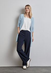 Ljusblå kavaj, vit t-shirt, mörkblå vida byxor och vita sneakers, mot en neutral bakgrund.