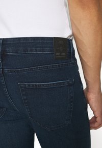 Tamno plave traperice s glatkom teksturom, koje imaju stražnji džep i crnu oznaku urezanu s "ONLY & SONS" na pojasu.