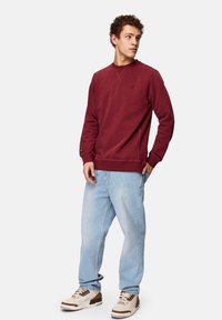 Maroon crew neck sweatshirt yang terbuat dari kain lembut, dipadukan dengan celana jeans biru muda. Detail mencolok termasuk manset dan hem yang diribbed. Sepatu sneaker netral.