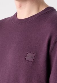 Gros plan d'une personne portant un pull en maille violet avec un col rond côtelé et un petit patch carré avec le logo « BOSS » sur la poitrine.