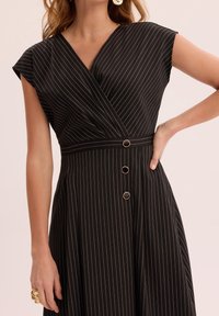 Femme portant une robe noire à fines rayures avec manches courtes et trois petits boutons ronds à la taille, main sur la hanche et bague en or à la main gauche.