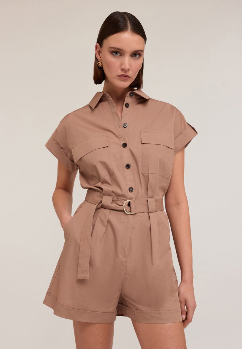 Jumpsuit corto marrone con maniche corte, dotato di due tasche frontali, vita con cintura e fibbia argentata, e chiusura con bottoni sul davanti. Tessuto con texture liscia.
