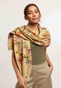 Écharpe florale en beige, ornée de diverses fleurs colorées. Texture douce avec des bords frangés. Drapée sur les épaules.