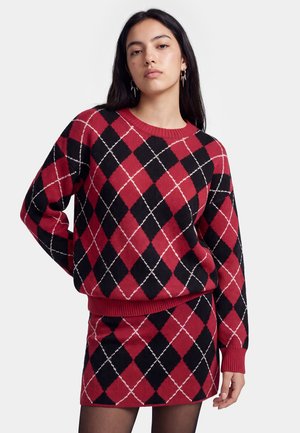 Jonge vrouw met lang donker haar draagt een rood-zwart argyle-trui en bijpassende rok, poserend tegen een effen lichte achtergrond.