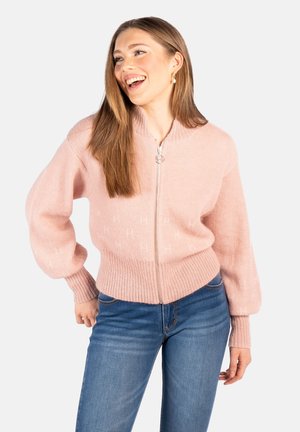 Smilende kvinne med langt brunt hår, iført perleøreringer, en rosa strikkegenser med glidelås og blå jeans, poserer med én hånd på hoften.