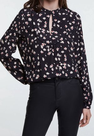 Femme portant un chemisier noir à imprimé floral blanc et rouge, avec une encolure goutte d'eau et des manches longues, associé à un pantalon noir ajusté.