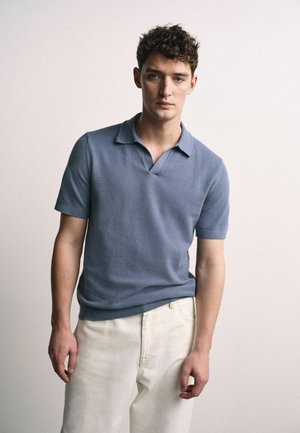 Jaunas vyras su garbanotais plaukais, dėvintis trumpomis rankovėmis mėlyną polo marškinėlį ir baltas kelnes, stovintis priešais vienodą šviesią fono spalvą.