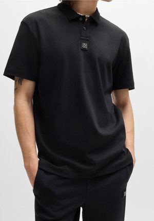 Man in een zwart polo-shirt met korte mouwen en bijpassende zwarte broek, voorzien van het HUGO-logo op zowel het shirt als de broek, met zijn handen in zijn zakken.