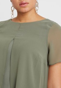 Olijfgroene blouse met een ronde halslijn, korte doorschijnende mouwen en een centrale verticale naaddetails. Lichtgewicht stof met een zachte textuur.