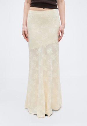 Kobieta ubrana w kremową, sięgającą podłogi koronkową spódnicę z motywem kwiatowym i dopasowaną talią, w połączeniu z krótkim ciemnym topem.