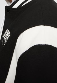 Černobílá varsity bunda s žebrovaným límcem, z vlněné směsi, a vyšitým logem na přední straně v blokových písmenech.