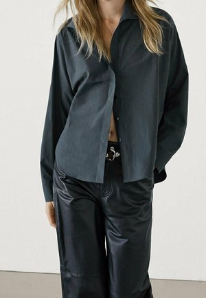 Personne portant une chemise gris foncé à boutons partiellement ouverte et un pantalon en cuir noir à jambes larges avec une ceinture noire.