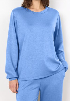 Kobieta ubrana w luźną, długorękawową, jasnoniebieską bluzkę i spodnie do kompletu, z jedną ręką w kieszeni.