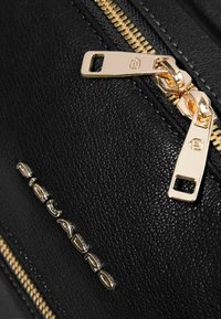 Zwarte leren tas met een gouden ritssluiting voorzien van een opvallende trekker en een geperst logo in metalen letters aan de zijkant.