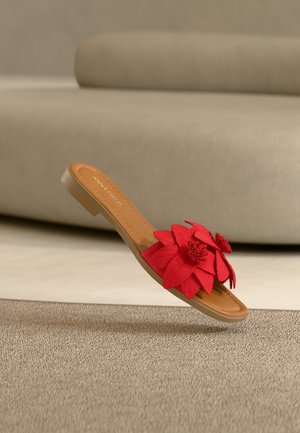 Sandalía plana de color canela con una gran flor de tela roja decorativa en la correa, colocada sobre un fondo de sofá beige y alfombra.