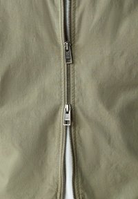 Nahaufnahme einer olivgrünen Jacke mit einem teilweise geöffneten silbernen Doppelreißverschluss, der ein weißes Kleidungsstück darunter enthüllt.