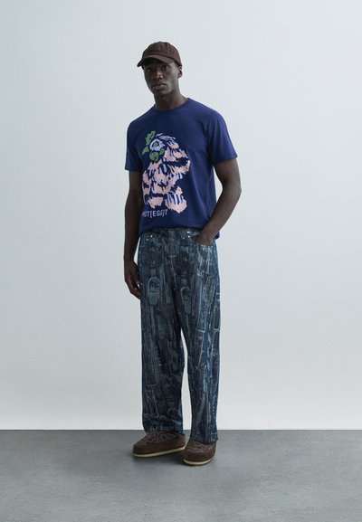 Μπλε ναυτικό T-shirt με λουλουδάτο σχέδιο, συνδυασμένο με χαλαρά παντελόνια σε σκούρο πλύσιμο με μοτίβο. Καφέ καπέλο και παπούτσια ολοκληρώνουν την εμφάνιση.