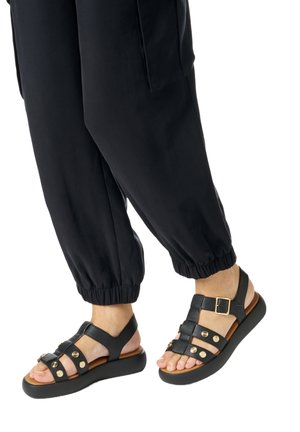 Pieds chaussés de sandales noires avec des clous dorés et des boucles, associés à un pantalon noir à poignets élastiques sur fond blanc.