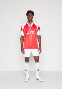 Camisola de futebol vermelha e branca com riscas verticais, apresentando um logotipo proeminente da JVC, acompanhada de calções brancos e meias até ao joelho com riscas pretas.