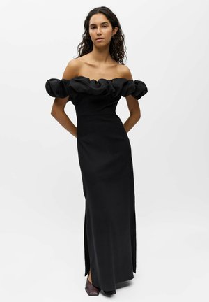 Frau trägt ein schwarzes, bodenlanges, schulterfreies Kleid mit gerüschtem Ausschnitt und steht mit den Händen hinter dem Rücken vor weißem Hintergrund.