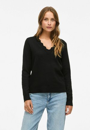 Schwarzer Strickpullover mit einem gewellten V-Ausschnitt und langen Ärmeln, kombiniert mit hellblauen Jeans. Glatte Textur und taillierte Passform.