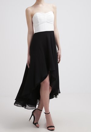 Trägerloses Kleid mit weißem, gerafftem Oberteil und asymmetrischem schwarzen Rock, kombiniert mit schwarzen High Heels mit Knöchelriemen vor neutralem Hintergrund.